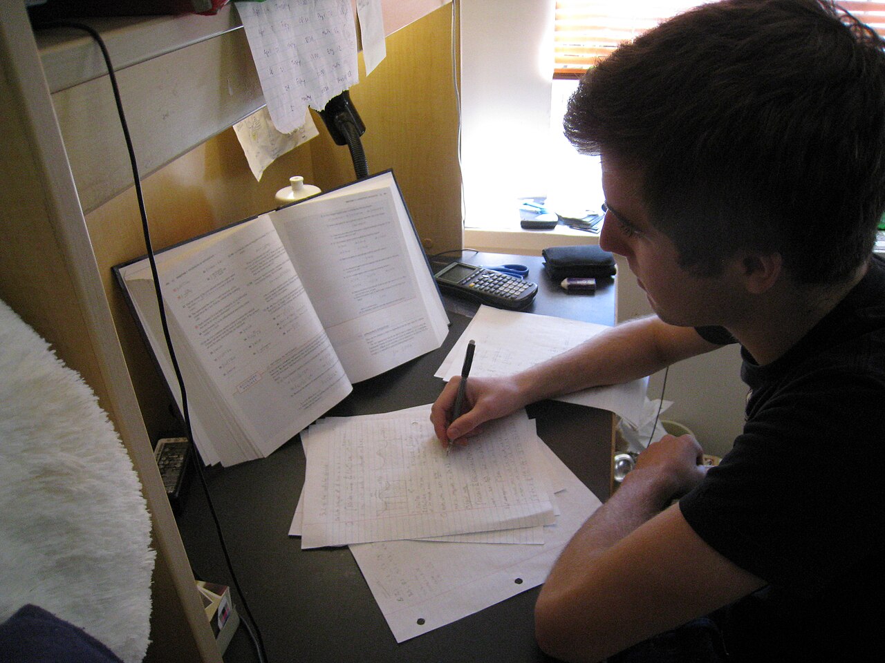 Un étudiant penché sur son bureau, en train d’écrire sur des feuilles à côté d’un livre ouvert